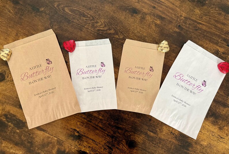 May include: Four paper favor bags with the text "A Little Butterfly is on the Way!" in a script font, with a butterfly graphic. Each bag has a different shade of brown or white. A red or gold foil-wrapped chocolate is attached to each bag.