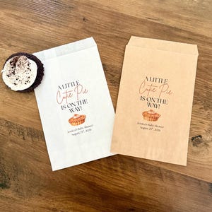 May include: Two rectangular paper favor bags, one white and one light brown, each printed with the text A LITTLE Cutie Pie IS ON THE WAY!, a small pie illustration, and the text Jessica's Baby Shower August 25th, 2026. A dark cookie sits on the white bag.