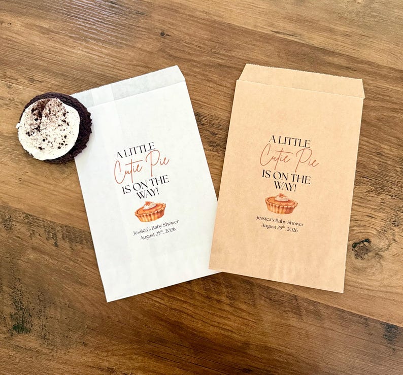 May include: Two rectangular paper favor bags, one white and one light brown, each printed with the text A LITTLE Cutie Pie IS ON THE WAY!, a small pie illustration, and the text Jessica's Baby Shower August 25th, 2026. A dark cookie sits on the white bag.