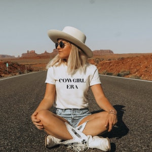 May include: A person wearing a white t-shirt with the text "COWGIRL ERA", a white cowboy hat, sunglasses, and denim shorts. The person is sitting on a road with a desert landscape in the background. The person is also wearing white cowboy boots.