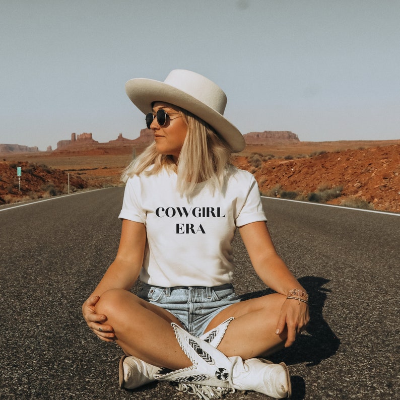 May include: A person wearing a white t-shirt with the text "COWGIRL ERA", a white cowboy hat, sunglasses, and denim shorts. The person is sitting on a road with a desert landscape in the background. The person is also wearing white cowboy boots.