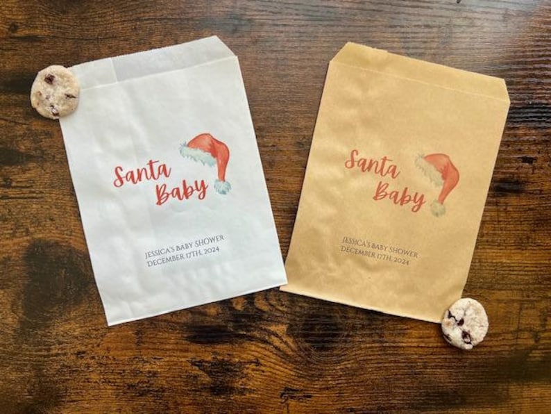 May include: Two paper treat bags, one white and one brown, with the text "Santa Baby" and a Santa hat graphic. The bags also include the text "JESSICA'S BABY SHOWER DECEMBER 17TH, 2024". A cookie rests near each bag.