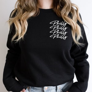 May include: Black crew neck sweatshirt with the word "Philly" written four times in a white cursive font. The sweatshirt is a solid black color and has a classic, casual style.