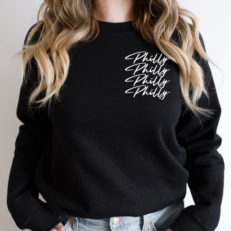 May include: Black crew neck sweatshirt with the word "Philly" written four times in a white cursive font. The sweatshirt is a solid black color and has a classic, casual style.