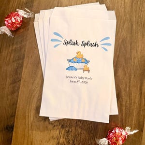Pode incluir: Uma pilha de sacos de papel branco com um gr&aacute;fico azul de uma banheira com patinhos de borracha amarelos e bolhas azuis. O texto diz Splish Splash, Jessica's Baby Bash e June 8th, 2026. Dois doces embrulhados em vermelho est&atilde;o pr&oacute;ximos.