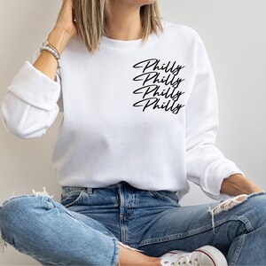 May include: White sweatshirt with the word "Philly" in a black script font repeated four times on the upper left chest. The sweatshirt is long-sleeved and worn by a person wearing blue jeans.