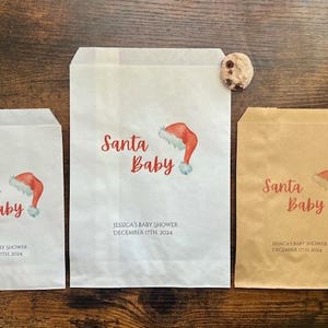 May include: Three paper gift bags with "Santa Baby" and a Santa hat illustration. The bags are white and brown, with "Jessica's Baby Shower December 17th 2024" printed. Chocolate chip biscuits are placed near the bags.