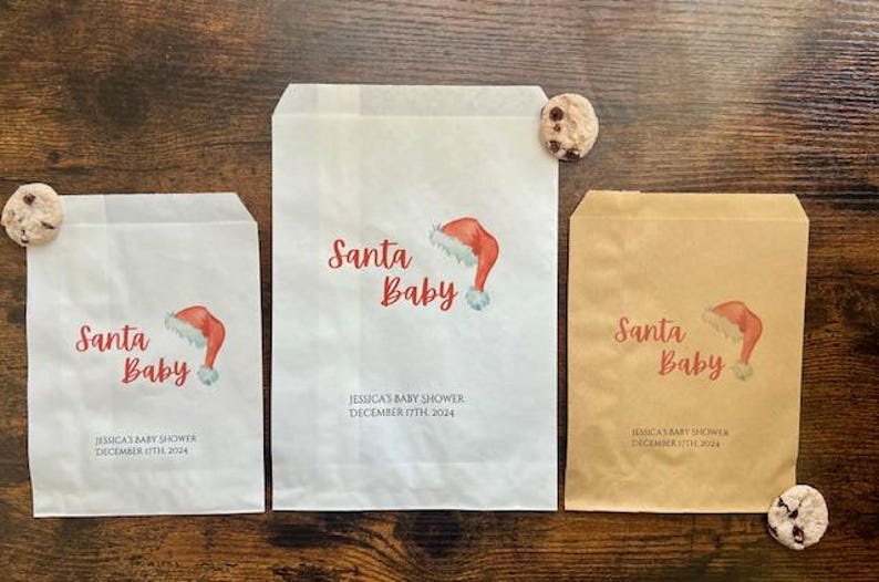 May include: Three paper gift bags with "Santa Baby" and a Santa hat illustration. The bags are white and brown, with "Jessica's Baby Shower December 17th 2024" printed. Chocolate chip biscuits are placed near the bags.