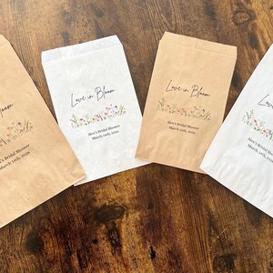 May include: Four paper favor bags, two in kraft brown and two in white, are arranged on a wooden surface. Each bag is printed with the words "Love in Bloom" and floral illustrations, along with event details.