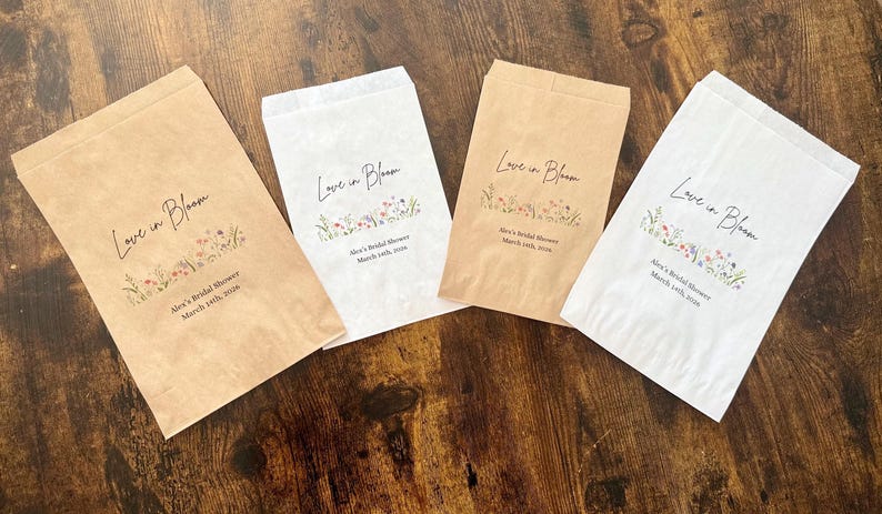 May include: Four paper favor bags, two in kraft brown and two in white, are arranged on a wooden surface. Each bag is printed with the words "Love in Bloom" and floral illustrations, along with event details.