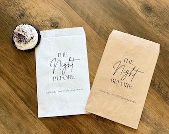 Puede incluir: Dos bolsas de papel rectangulares, una blanca y otra marrón, impresas con el texto negro THE Night BEFORE y Jessica & Dave's Rehearsal Dinner. Una pequeña galleta redonda de chocolate negro con glaseado blanco se encuentra junto a la bolsa blanca.