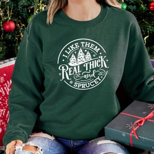 Pu&ograve; includere: Felpa verde scuro con grafica bianca circolare con la scritta "I LIKE THEM REAL THICK and SPRUCEY", con alberi di pino e fiocchi di neve. La felpa &egrave; a tinta unita e ha un girocollo.