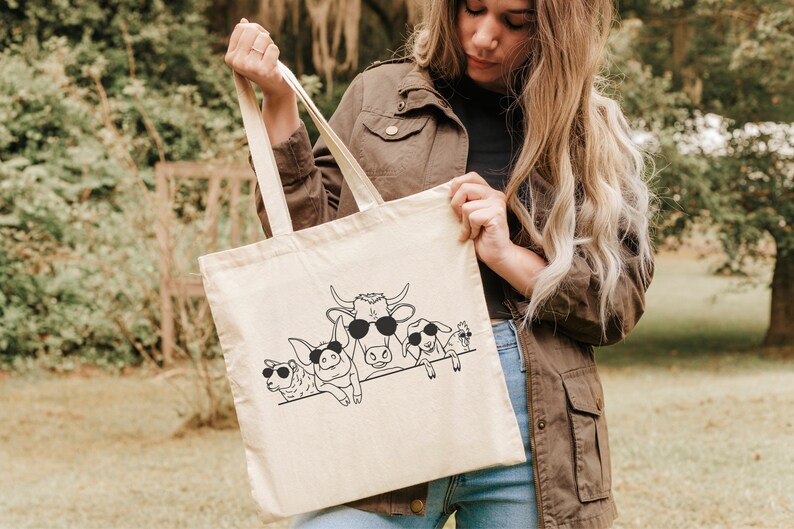 May include: A beige canvas tote bag featuring a black line drawing of farm animals wearing sunglasses. The animals include a cow, sheep, and pig. The bag is held by a person outdoors.