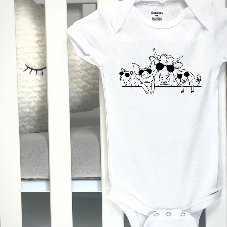 Puede incluir: Body de beb&eacute; blanco con un dibujo negro de una vaca, un cerdo, una oveja y una cabra con gafas de sol. La prenda tiene mangas cortas y cierre de broche. Etiqueta de la marca Gerber.