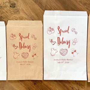 May include: Four rectangular paper bags in white and brown kraft paper, featuring red printed graphics of baby items, pizza, and wine glasses. The text reads Special Delivery, Jessica's Baby Shower, and July 15th, 2026, with two oval cookies.