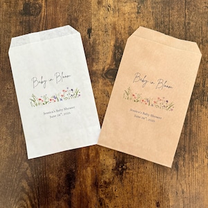 以下が含まれることがあります： エレガントな筆記体で「Baby in Bloom」と書かれた、白とタンの2つの紙製のギフトバッグ。テキストの下には、カラフルな花柄のデザインがあります。「Jessica's Baby Shower June 28, 2025」の文字が下に印刷されています。