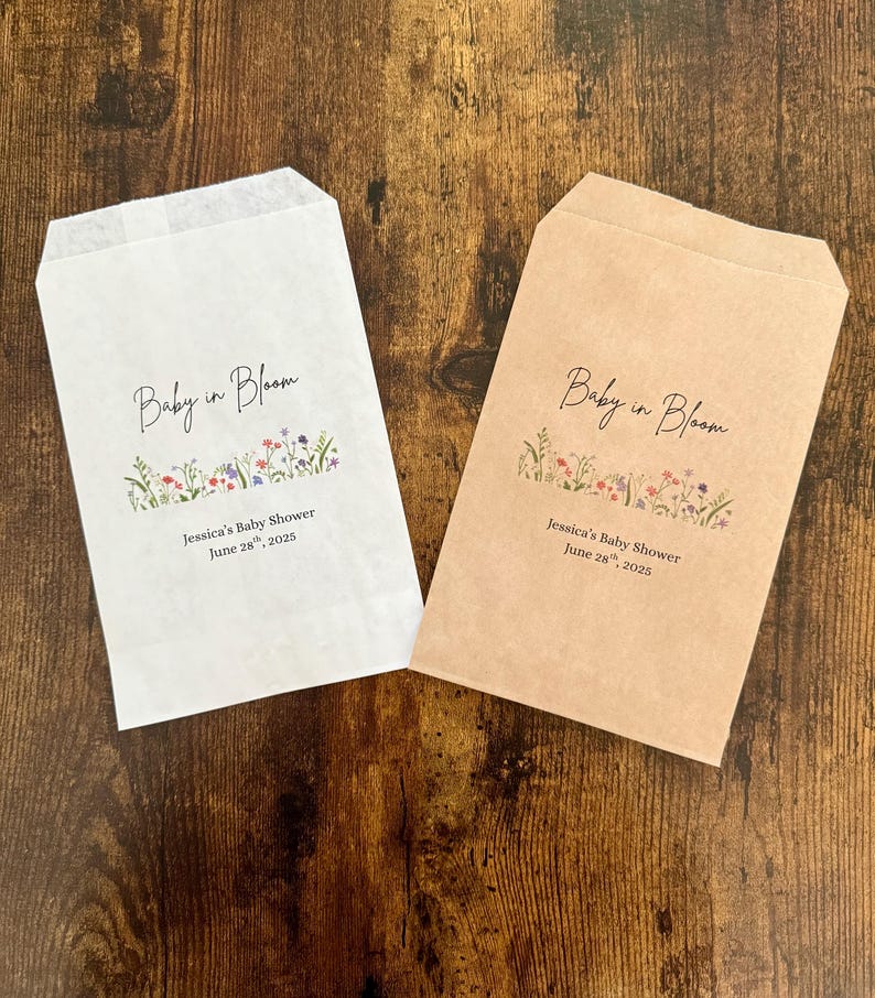 以下が含まれることがあります： エレガントな筆記体で「Baby in Bloom」と書かれた、白とタンの2つの紙製のギフトバッグ。テキストの下には、カラフルな花柄のデザインがあります。「Jessica's Baby Shower June 28, 2025」の文字が下に印刷されています。