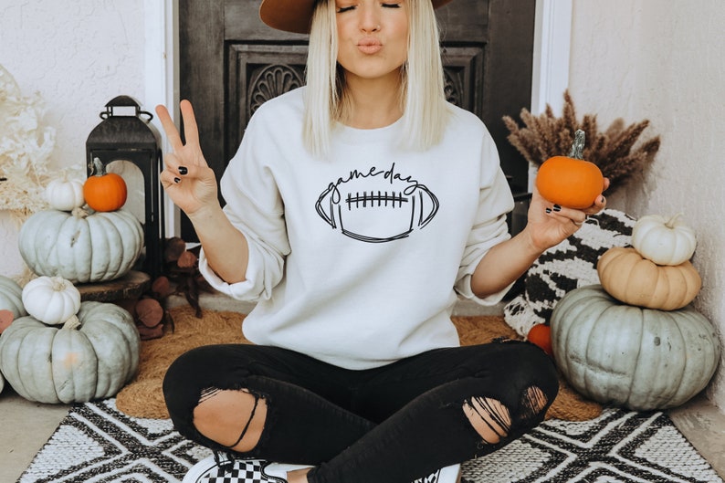 Puede incluir: Sudadera color crema con el texto "gameday" sobre un gr&aacute;fico de f&uacute;tbol americano. La persona sostiene una calabaza naranja y hace el signo de la paz. En el fondo hay calabazas decorativas y decoraci&oacute;n oto&ntilde;al.