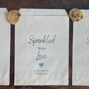 May include: Three white paper treat bags with the text "Sprinkled With Love" and a heart graphic in pink, blue, and black. Each bag has a chocolate chip cookie attached. The text "Jessica's Sprinkle, March 9th 2024" is printed below the heart.