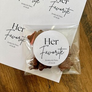 Peut inclure: Un sachet en plastique transparent contenant des petits biscuits bruns, scell&eacute; par un autocollant blanc rond avec le texte noir "Her Favorite" suivi de "Jessica & Dave" et la date "07.14.26". D'autres autocollants identiques sont sur une feuille.