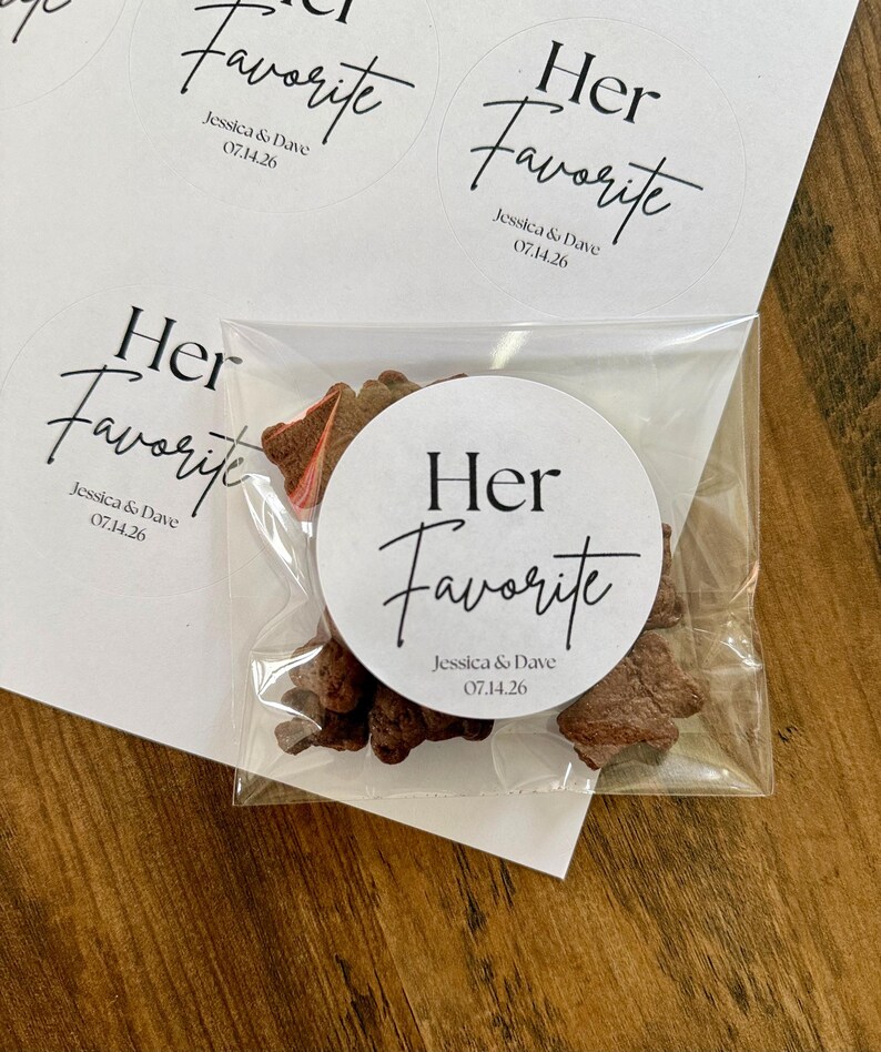 Peut inclure: Un sachet en plastique transparent contenant des petits biscuits bruns, scell&eacute; par un autocollant blanc rond avec le texte noir "Her Favorite" suivi de "Jessica & Dave" et la date "07.14.26". D'autres autocollants identiques sont sur une feuille.