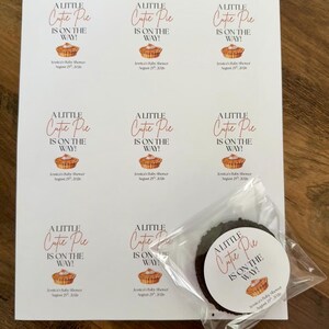 Op de afbeelding: Een wit vel met acht identieke rechthoekige stickers met de tekst A LITTLE Cutie Pie IS ON THE WAY! en een illustratie van een taartje, plus Jessica's Baby Shower August 25th, 2026. E&eacute;n ronde sticker zit in een doorzichtig zakje.