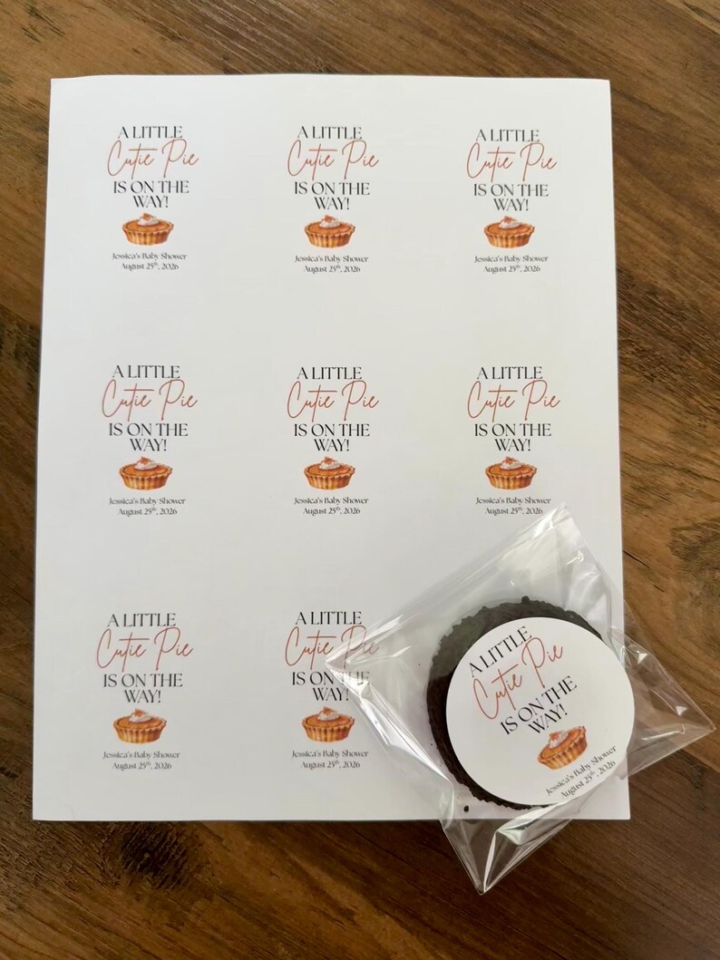 Op de afbeelding: Een wit vel met acht identieke rechthoekige stickers met de tekst A LITTLE Cutie Pie IS ON THE WAY! en een illustratie van een taartje, plus Jessica's Baby Shower August 25th, 2026. E&eacute;n ronde sticker zit in een doorzichtig zakje.