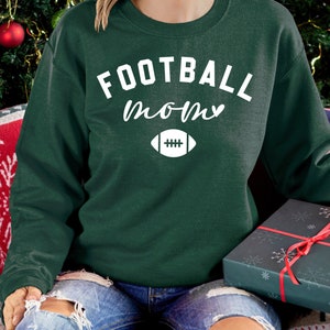 May include: A dark green sweatshirt with the words "FOOTBALL mom" in white script and a white football graphic. The sweatshirt is part of a holiday theme, with a gift box and Christmas decorations in the background.