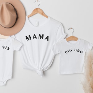 May include: White t-shirts and a onesie with black text that reads "MAMA", "BIG BRO", and "LIL SIS". A tan hat hangs above the shirts. The shirts are on wooden hangers. A sprig of dried pampas grass is on the right.