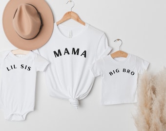 May include: White t-shirts and a onesie with black text that reads "MAMA", "BIG BRO", and "LIL SIS". A tan hat hangs above the shirts. The shirts are on wooden hangers. A sprig of dried pampas grass is on the right.