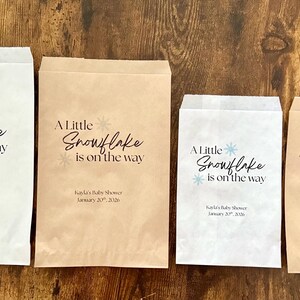 May include: Four paper treat bags with the text "A Little Snowflake is on the way" in black and blue. The bags are white and brown, with a snowflake graphic. The text "Kayla's Baby Shower January 20, 2026" is also printed on the bags.
