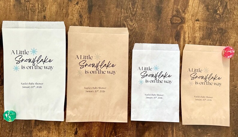 May include: Four paper treat bags with the text "A Little Snowflake is on the way" in black and blue. The bags are white and brown, with a snowflake graphic. The text "Kayla's Baby Shower January 20, 2026" is also printed on the bags.