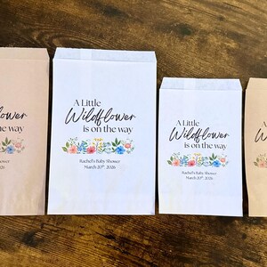 May include: Four paper favor bags with the text "A Little Wildflower is on the way" and floral illustrations. The bags are in various sizes and colors, including white and light brown. Text at the bottom reads "Rachel's Baby Shower March 20, 2026."