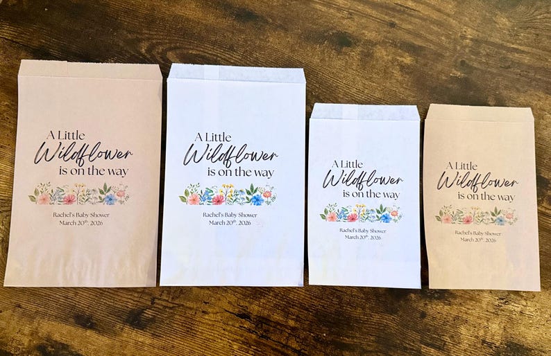 May include: Four paper favor bags with the text "A Little Wildflower is on the way" and floral illustrations. The bags are in various sizes and colors, including white and light brown. Text at the bottom reads "Rachel's Baby Shower March 20, 2026."
