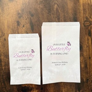 Puede incluir: Dos bolsas de papel blancas con el texto "OUR LITTLE Butterfly IS TURNING ONE!" en escritura morada y un gr&aacute;fico de mariposa. El texto "Jessica's First Birthday April 22, 2026" tambi&eacute;n est&aacute; impreso en las bolsas.