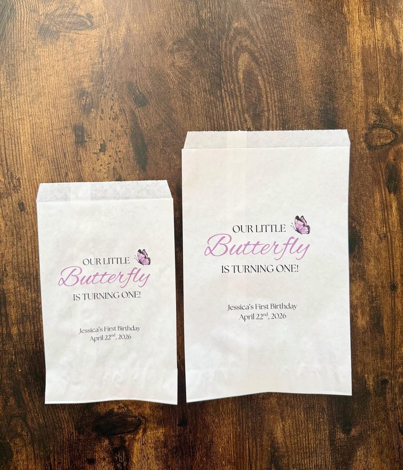 Puede incluir: Dos bolsas de papel blancas con el texto "OUR LITTLE Butterfly IS TURNING ONE!" en escritura morada y un gr&aacute;fico de mariposa. El texto "Jessica's First Birthday April 22, 2026" tambi&eacute;n est&aacute; impreso en las bolsas.