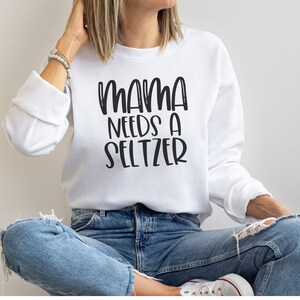 Puede incluir: Sudadera blanca con el texto "MAMA NEEDS A SELTZER" en letras negras y en negrita. La sudadera tiene mangas largas y cuello redondo. La persona de la imagen lleva vaqueros azules.