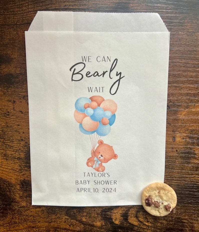 May include: A white paper bag with the text "We Can Bearly Wait" and a watercolor illustration of a teddy bear holding balloons. The bag is for Taylor's Baby Shower, April 10, 2024. A chocolate chip cookie rests on the surface.