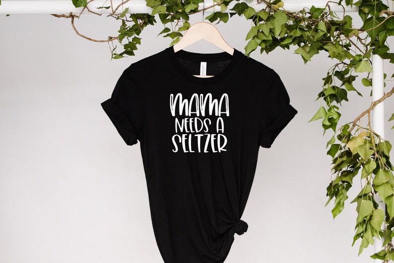 May include: Black t-shirt with the white text "MAMA NEEDS A SELTZER". The shirt is hanging on a wooden hanger against a white background with green leaves.