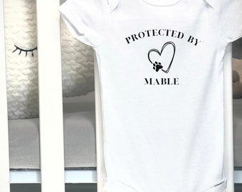 May include: White baby onesie with the text "PROTECTED BY MABLE" in black. The design includes a heart with a paw print inside. The onesie is made of soft material and has snap closures.