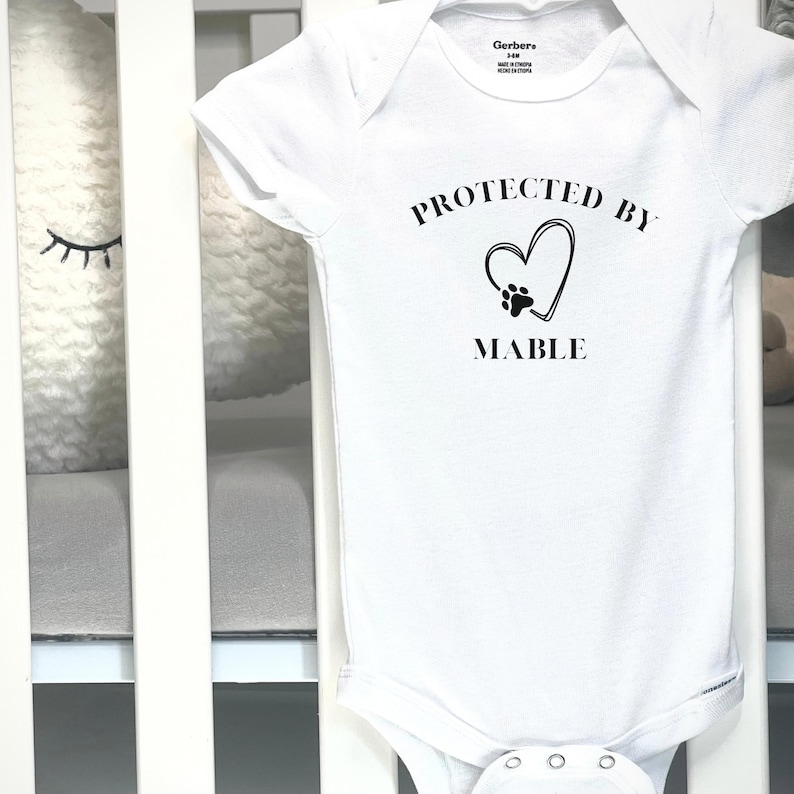 May include: White baby onesie with the text "PROTECTED BY MABLE" in black. The design includes a heart with a paw print inside. The onesie is made of soft material and has snap closures.