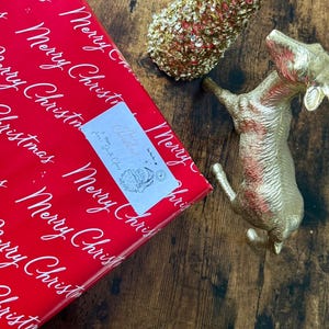 May include: A Christmas-themed image with a red gift box printed with white "Merry Christmas" text, a gold dog ornament, and a gold Christmas tree decoration. A small label on the gift box reads "Merry Christmas! From Santa Claus".