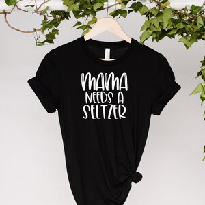 Puede incluir: Camiseta negra con el texto blanco "MAMA NEEDS A SELTZER". La camiseta est&aacute; colgada en una percha de madera sobre un fondo blanco con hojas verdes.
