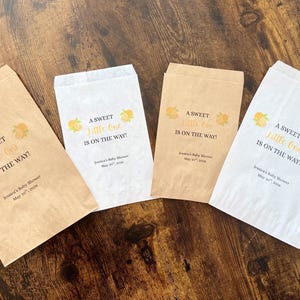 K&ouml;nnte beinhalten: Vier Papier-Gastgeschenkt&uuml;ten mit dem Text "A Sweet Little One Is On The Way!" in Schwarz und Gold. Jede T&uuml;te hat ein gelbes Blumendesign und das Datum der Babyparty. Die T&uuml;ten sind in Braun- und Wei&szlig;t&ouml;nen gehalten.