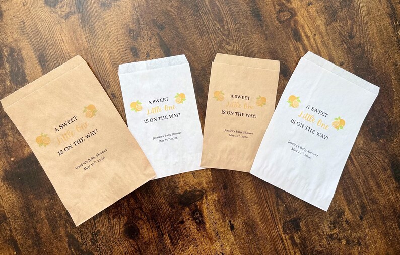 K&ouml;nnte beinhalten: Vier Papier-Gastgeschenkt&uuml;ten mit dem Text "A Sweet Little One Is On The Way!" in Schwarz und Gold. Jede T&uuml;te hat ein gelbes Blumendesign und das Datum der Babyparty. Die T&uuml;ten sind in Braun- und Wei&szlig;t&ouml;nen gehalten.
