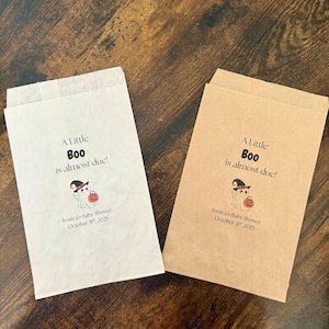 K&ouml;nnte beinhalten: Zwei Papiert&uuml;ten, eine wei&szlig;e und eine beige, mit dem Text "A Little Boo is almost due!" und einer Geistergrafik. Die T&uuml;ten sind f&uuml;r Jessicas Babyparty, 31. Oktober 2025.