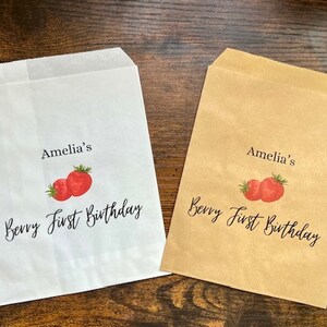 May include: Two paper treat bags, one white and one tan, with a strawberry design. The bags are printed with the text "Amelia's Berry First Birthday" in black script, perfect for a first birthday party.