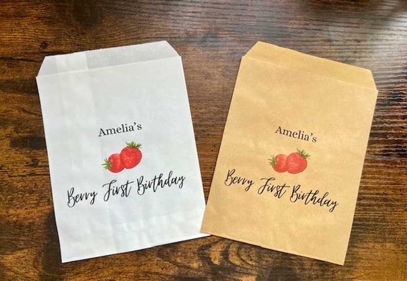 May include: Two paper treat bags, one white and one tan, with a strawberry design. The bags are printed with the text "Amelia's Berry First Birthday" in black script, perfect for a first birthday party.