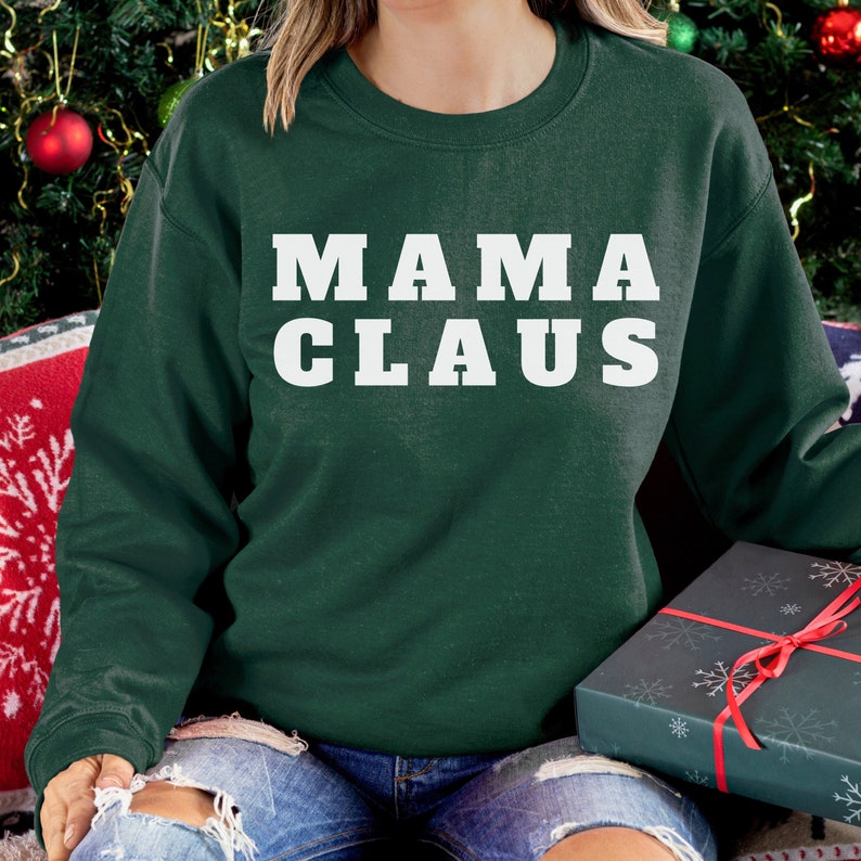 Op de afbeelding: Een donkergroene sweatshirt met de woorden "MAMA CLAUS" in dikke witte letters. De sweatshirt maakt deel uit van een collectie met kerstthema. Een ingepakte geschenkdoos met een rood lint is zichtbaar.