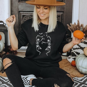 May include: Black sweatshirt featuring a white graphic of dancing skeletons and bats. The sweatshirt is part of a Halloween-themed collection. The image also includes pumpkins and decorative elements.
